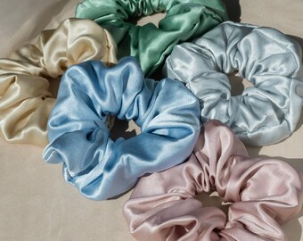 May include: A collection of satin scrunchies in various pastel colors, including light blue, pink, green, and cream. The scrunchies are arranged in a circular pattern, showcasing their soft, gathered fabric. These hair accessories add a touch of elegance.