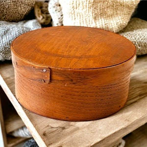 May include: A round wooden box with a natural wood finish. The box has a lid and is made of wood with a grain pattern. The box is sitting on a wooden shelf.