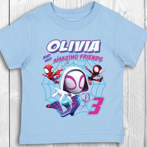 May include: Light blue t-shirt featuring a graphic of Spider-Gwen and two other Spider-Man characters. The text "Olivia and her amazing friends" is printed on the shirt. The number "3" is also printed on the shirt.