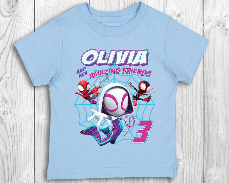 May include: Light blue t-shirt featuring a graphic of Spider-Gwen and two other Spider-Man characters. The text "Olivia and her amazing friends" is printed on the shirt. The number "3" is also printed on the shirt.