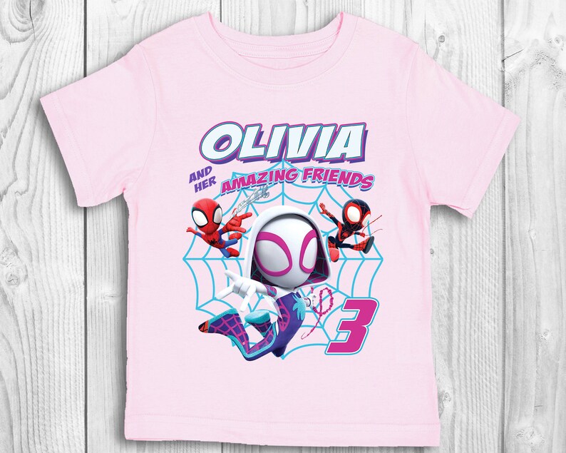 May include: Pink t-shirt featuring a graphic of Spider-Gwen and two other Spider-Man characters. The text "Olivia and her amazing friends" is above the graphic and the number "3" is below the graphic.