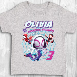 May include: Grey t-shirt featuring a Spider-Gwen character design. The shirt includes the text "Olivia and her Amazing Friends" and the number "3".