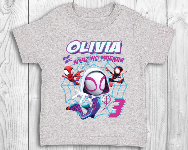May include: Grey t-shirt featuring a Spider-Gwen character design. The shirt includes the text "Olivia and her Amazing Friends" and the number "3".