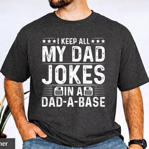 May include: Dark heather t-shirt with the text "I KEEP ALL MY DAD JOKES IN A DAD-A-BASE" in white. The design includes stars and a floppy disk graphic. The shirt is a casual wear item.