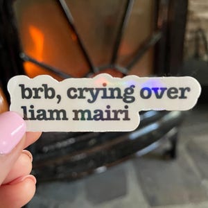 Puede incluir: Una pegatina blanca con letras iridiscentes que dice "brb, crying over liam mairi".