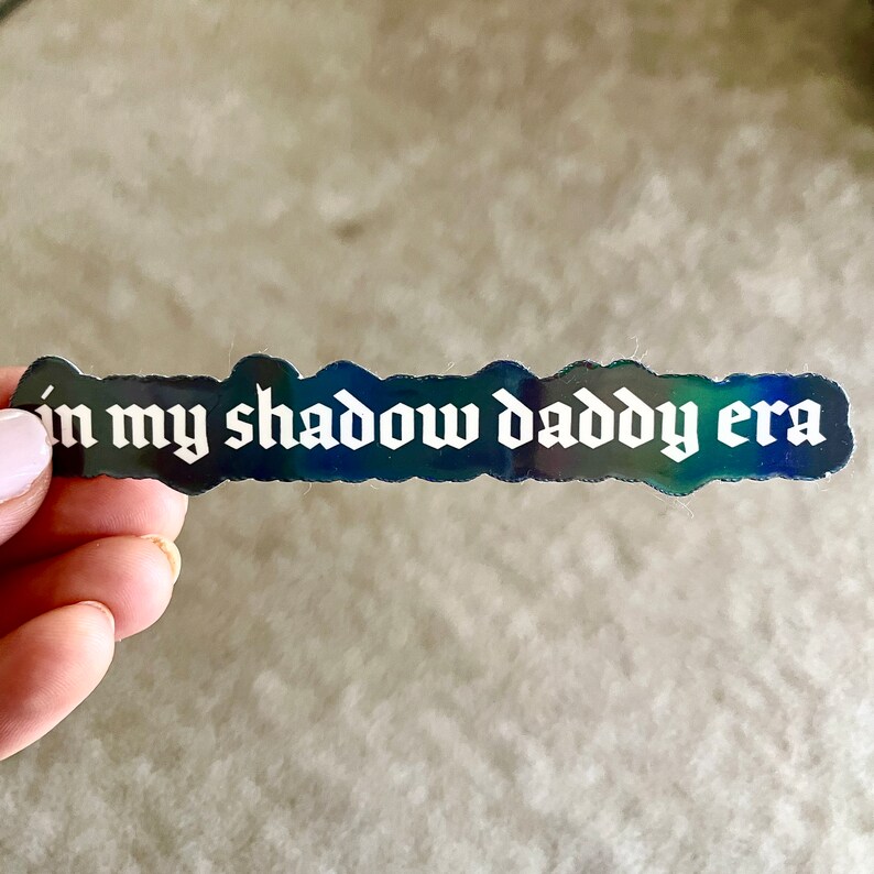 May include: A black and iridescent sticker with the text "in my shadow daddy era" in white gothic font.