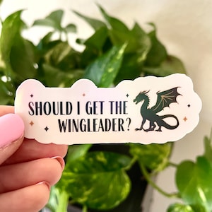 Puede incluir: Una pegatina blanca con un dragón verde y el texto "Should I get the Wingleader?" en negro. La pegatina tiene un acabado holográfico y detalles de purpurina dorada.