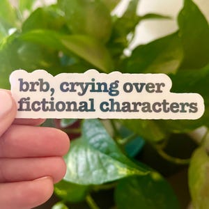 Puede incluir: Una pegatina blanca con el texto "brb, crying over fictional characters" en negro.