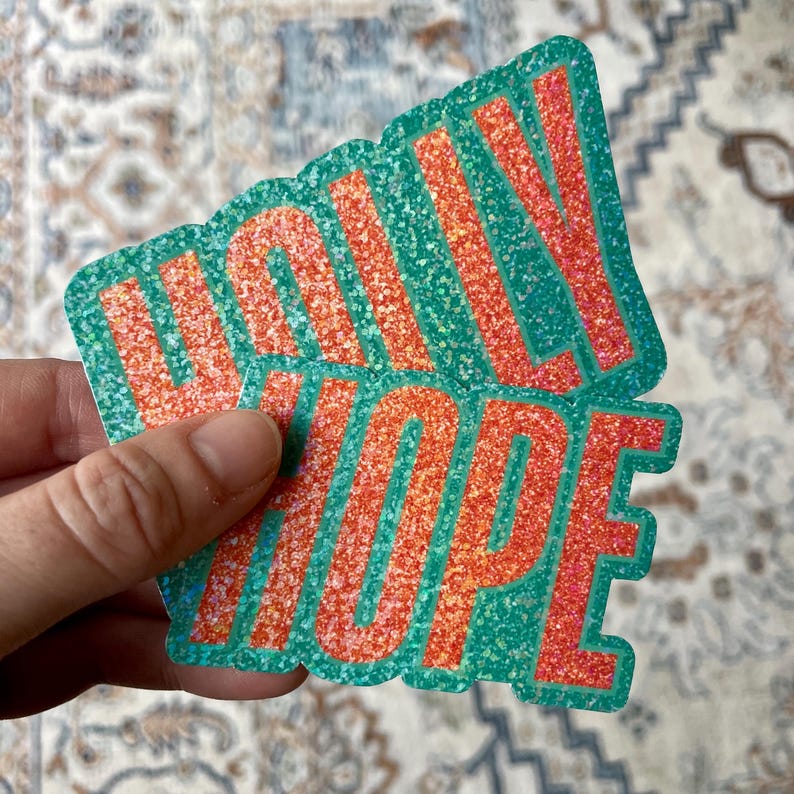 May include: Two glittery stickers with the words "WALLY HOPE" in orange letters on a teal background. The stickers are held in a hand, and the background is a patterned rug.