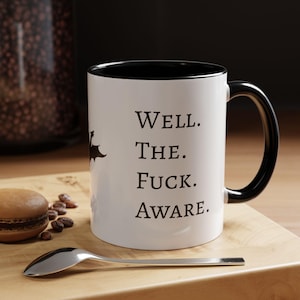 Pode incluir: Caneca de cerâmica branca com interior e alça pretos. A caneca apresenta o texto "Well. The. Fuck. Aware." em preto, juntamente com uma silhueta de morcego. Uma colher e um macaron estão sobre uma superfície de madeira.