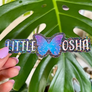 Può includere: Un adesivo olografico con sfondo nero glitterato e una farfalla blu con ali iridescenti. L'adesivo dice "Little Osha" in lettere bianche.