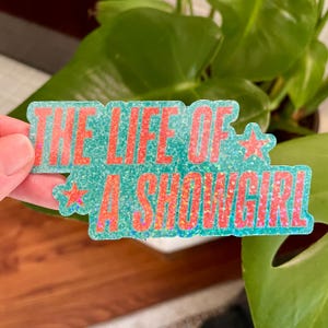 May include: A teal glitter sticker with the words "THE LIFE OF A SHOWGIRL" in red, with a holographic shimmer outline. The sticker features a star on each end of the text. It is held in front of a green plant.