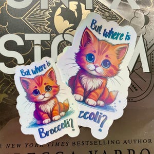 May include: Two iridescent stickers featuring a cartoon orange tabby cat with blue eyes. The text on the stickers reads "But where is Broccoli?"