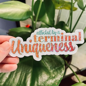 May include: A sticker with the phrase "afflicted by a terminal Uniqueness" in orange glitter lettering. The sticker has a white border and is held in front of a background of green leaves.