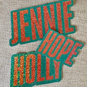 May include: Three glittery name cutouts: "JENNIE", "HOPE", and "HOLLY". The names are in a bold, orange font with a green outline. The glitter adds a sparkling effect to the names, making them stand out.