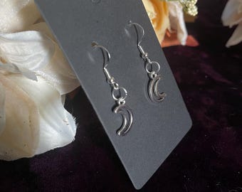 May include: Silver-toned crescent moon earrings displayed on a black earring card. The earrings have a simple, elegant design with a curved moon shape. The background features floral accents and a dark purple velvet surface.