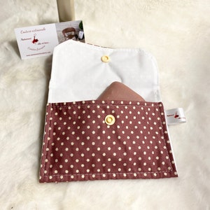 Peut inclure: Une petite pochette en tissu à pois marrons et blancs avec une doublure blanche et un bouton jaune. La pochette est ouverte et un rectangle brun est à l'intérieur.