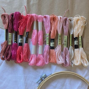 May include: An assortment of embroidery threads in shades of pink, peach, and cream, displayed on a neutral background. The threads are bundled with paper labels. A wooden embroidery hoop and white fabric are also visible, indicating a craft project.