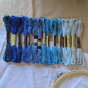 May include: A collection of blue embroidery threads in diverse tones, displayed on white fabric. The threads are labelled with black and yellow tags. A wooden embroidery hoop is in the bottom right, indicating a crafting project.