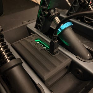 Puede incluir: Primer plano de un dispositivo rectangular negro con un cable USB conectado, posicionado dentro de un conjunto mecánico gris oscuro. El dispositivo tiene una superficie acanalada y una luz verde. El conjunto incluye componentes negros y un acento azul.