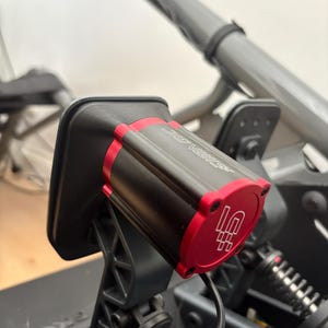 Op de afbeelding: Close-up van een zwart en rood elektronisch apparaat met de tekst "SIMAGIC" en een logo. Het apparaat is gemonteerd op een zwarte beugel en aangesloten op een blauwe kabel. De achtergrond bevat een grijs metalen frame.