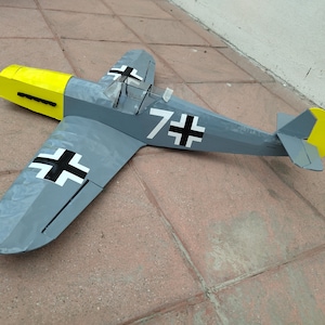 Op de afbeelding: Een grijs en geel modelvliegtuig met zwart-witte markeringen, waaronder het nummer "7+" en een kruissymbool. Het speelgoedvliegtuig ligt op een bakstenen oppervlak.