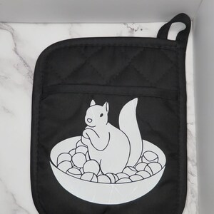 Könnte beinhalten: Schwarzer, gesteppter Topflappen mit einer weißen Grafik eines Eichhörnchens in einer Schüssel mit runden Objekten. Der Topflappen hat eine Schlaufe zum Aufhängen.