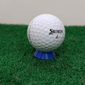 May include: A white Srixon golf ball, marked with the number 4, sits atop a blue tee on a green surface. The golf ball has a dimpled surface. The Srixon logo is printed in black.