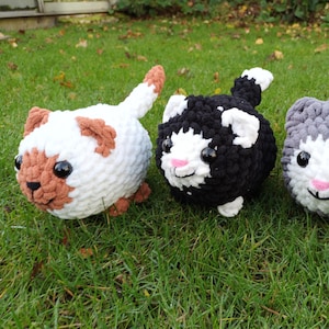 May include: Three crocheted cat plushies in different colours: white with brown accents, black with white accents, and grey with white accents. The plushies have large eyes and a smiling expression.