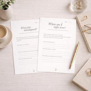 Op de afbeelding: Twee witte journaalpagina's met de tekst "What feels misaligned?" en "Where am I right now?" worden weergegeven. Een gouden pen ligt op de rechterpagina. De pagina's liggen op een lichtgekleurd oppervlak, met een kopje koffie en kleine decoratieve items.