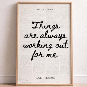Op de afbeelding: Een ingelijste print met een licht houten frame. De print toont de tekst "Things are always working out for me" in zwart schrift op een achtergrond van vervaagde tekst. De woorden "Daily Reminder" en "A Gentle Note" zijn ook gedrukt.