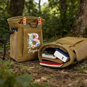 May include: Two brown canvas pouches with drawstring closures. One pouch features a floral "B" initial and the name "Brayder". The other pouch is open, revealing small electronics and wallets. A mesh pocket and orange and white accessories are visible.