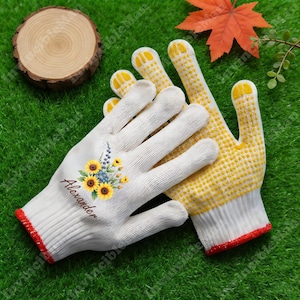 May include: White gardening gloves with a red trim and yellow dotted palms. One glove features a floral design with sunflowers and the name "Alexander". The gloves are placed on green grass.