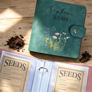 Op de afbeelding: Een zeegroen zaadboek met de naam "Sophia's" en de woorden "SEED BOOK" op de kaft. De kaft heeft een bloemenontwerp. Het boek is open en toont pagina's met zaadinfo-kaarten. Een hoop aarde ligt naast het boek.