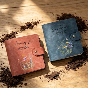 May include: Two seed books, one in a rust hue and the other in blue, featuring floral illustrations and the names "Henry's" and "Sophia's" on the covers. Each book has a snap closure. The books are on a wooden surface with soil.