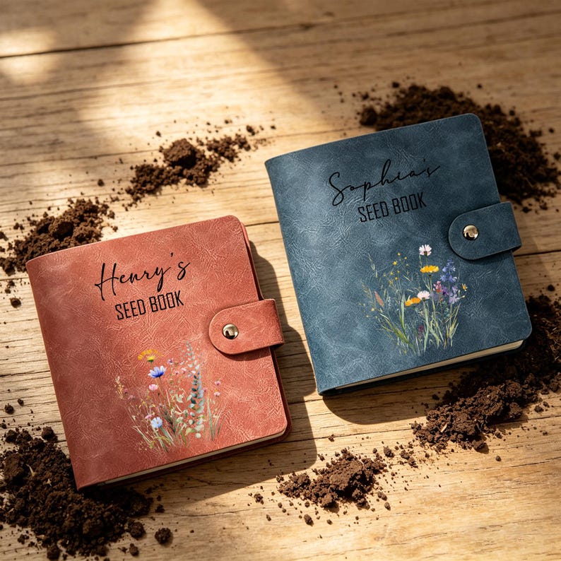 May include: Two seed books, one in a rust hue and the other in blue, featuring floral illustrations and the names "Henry's" and "Sophia's" on the covers. Each book has a snap closure. The books are on a wooden surface with soil.