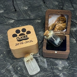 May include: A wooden box with a paw print and the name "Claire" engraved on the lid. The box is open and contains a small glass jar with a cork stopper. The jar contains a white feather. The box also has the dates "2016-2024" engraved on the lid.