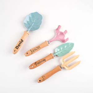 May include: Four small garden tools with wooden handles and pastel-coloured heads. The tools include a leaf-shaped trowel, a three-pronged cultivator, a leaf-shaped scoop, and a three-pronged fork. The handles are engraved with names.