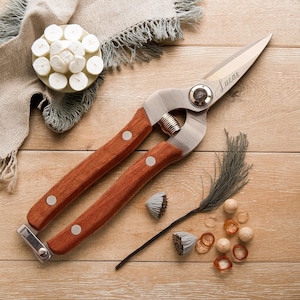 May include: A pair of gardening shears with wooden handles and silver blades, engraved with the name "Lucas". The shears are next to a small arrangement of dried flowers and decorative elements, all on a wooden surface.