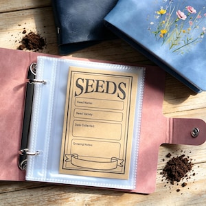 May include: Pink binder with clear sleeves holding a seed packet. The tan seed packet has the word "SEEDS" in bold letters. Spaces are provided for seed name, variety, date collected, and growing notes. Other blue binders are in the background. Soil is scattered around.