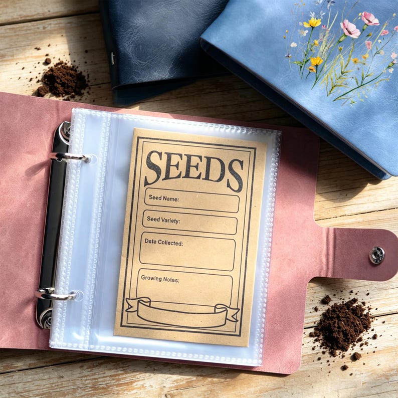May include: Pink binder with clear sleeves holding a seed packet. The tan seed packet has the word "SEEDS" in bold letters. Spaces are provided for seed name, variety, date collected, and growing notes. Other blue binders are in the background. Soil is scattered around.