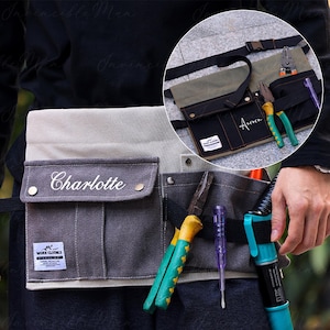 May include: A canvas work apron in grey and beige with multiple pockets. The apron is personalised with the name "Charlotte". Tools, including pliers and a screwdriver, are visible.