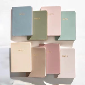 May include: A collection of eight notebooks in various pastel colors, including blue, green, pink, and beige. Each notebook has the word "NOTES" in gold lettering on the cover. The notebooks are arranged on a white surface.
