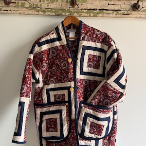 May include: Quilted jacket with a floral pattern in red, cream, and navy blue. The jacket has a square block design, wooden buttons, and two front pockets. It is hanging on a wooden hanger. The jacket's length is approximately 76 cm.