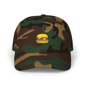 May include: Camouflage baseball cap with an embroidered cartoon hamburger. The cap features a classic six-panel design with a curved brim. The hamburger is yellow, red, and green, adding a pop of color against the brown, green, and tan camouflage pattern.