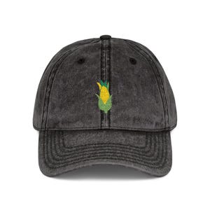 Op de afbeelding: Een donkergrijze baseballcap met een gebogen klep. De cap heeft een geborduurde maïsafbeelding in geel, groen en wit op de voorkant. De hoed heeft een vintage, versleten uitstraling.