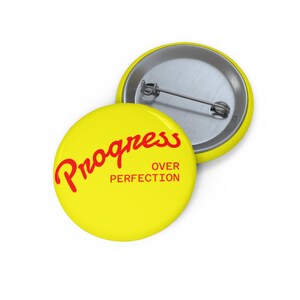 Op de afbeelding: Twee gele ronde buttons met de tekst "Progress Over Perfection" in het rood. Eén button staat in de focus, terwijl de andere gedeeltelijk zichtbaar is op de achtergrond. De buttons hebben een metalen pin achterkant.