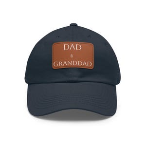 Op de afbeelding: Marineblauwe baseballpet met een bruine leren patch. De patch toont de witte woorden "DAD & GRANDDAD". De pet heeft een klassiek zes-panelen ontwerp met een gebogen klep.