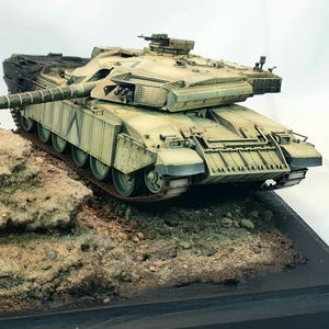 Könnte beinhalten: Ein detailliertes Modell eines Militärpanzers, bemalt in Wüsten-Tarnfarben. Der Panzer steht auf einer strukturierten, sandigen Basis, montiert auf einer schwarzen rechteckigen Plattform. Der lange Kanonenlauf erstreckt sich nach vorne, mit feinen Details.