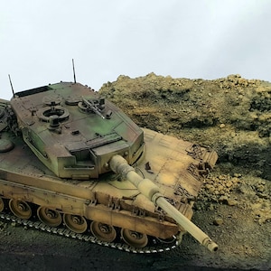 Puede incluir: Maqueta detallada de un tanque con un patrón de camuflaje verde, marrón y beige. El tanque tiene un cañón largo y varias ruedas. El fondo presenta un terreno rocoso y un cielo nublado, lo que sugiere un tema militar o histórico.